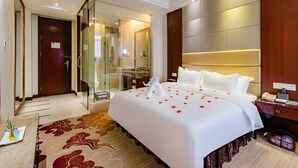 Habitación de hotel con una cama grande, ducha con cerramiento de vidrio, cabecero de madera y mesita de noche con lámpara.