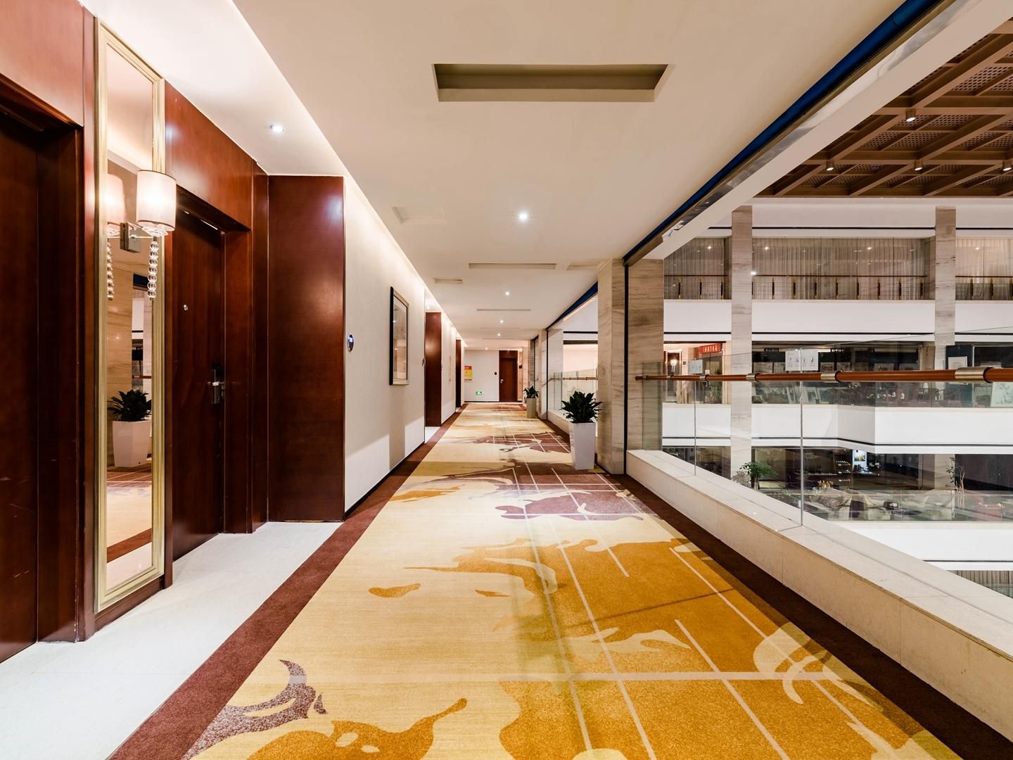 Pasillo de hotel con puertas de madera, alfombra estampada y vista del nivel inferior a través de una barandilla de cristal.