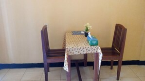 A small room with a table covered by a floral tablecloth, two wooden chairs, a vase with flowers, and a box labeled 'PASEO'. - OYO 91230 Kelimutu Eco Village (Moni)