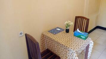 Une petite table à manger recouverte d’une nappe à motifs floraux, deux chaises en bois, un vase contenant une seule fleur et une boîte de mouchoirs.
