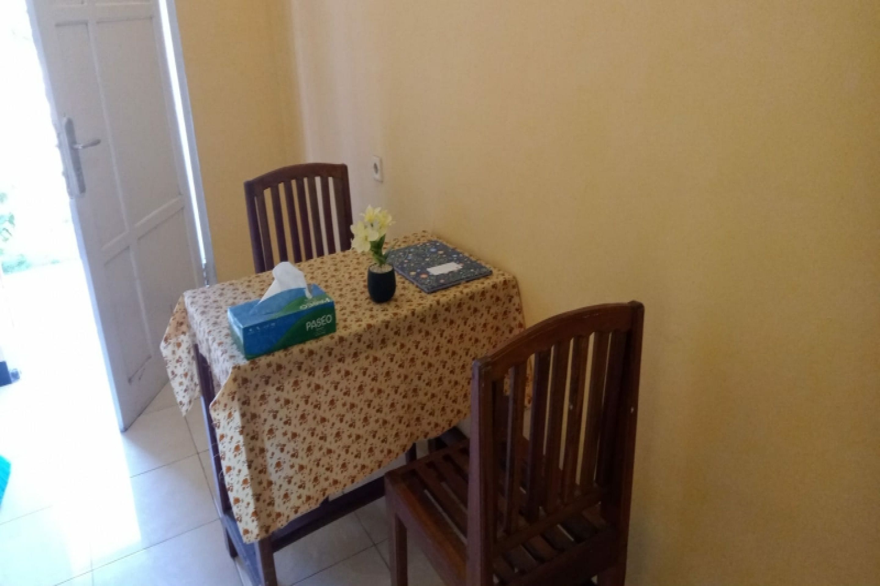 A dining area with a table covered by a floral tablecloth, two wooden chairs, a vase with flowers, and a box of tissues.