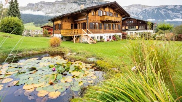 Una cabaña de madera con balcón, un estanque cubierto de lirios y montañas al fondo.