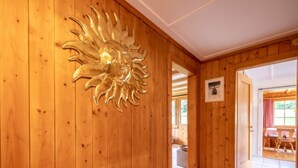 Un pasillo interior de madera con una decoración mural en forma de sol, un cuadro enmarcado y una vista de una habitación contigua con una mesa y sillas.