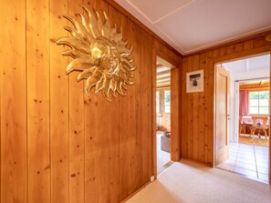 A wooden interior hallway with a sun-shaped wall decoration, a framed picture, and a glimpse of an adjacent room with a table and chairs. - Chalet Cauma by Interhome (Flims)