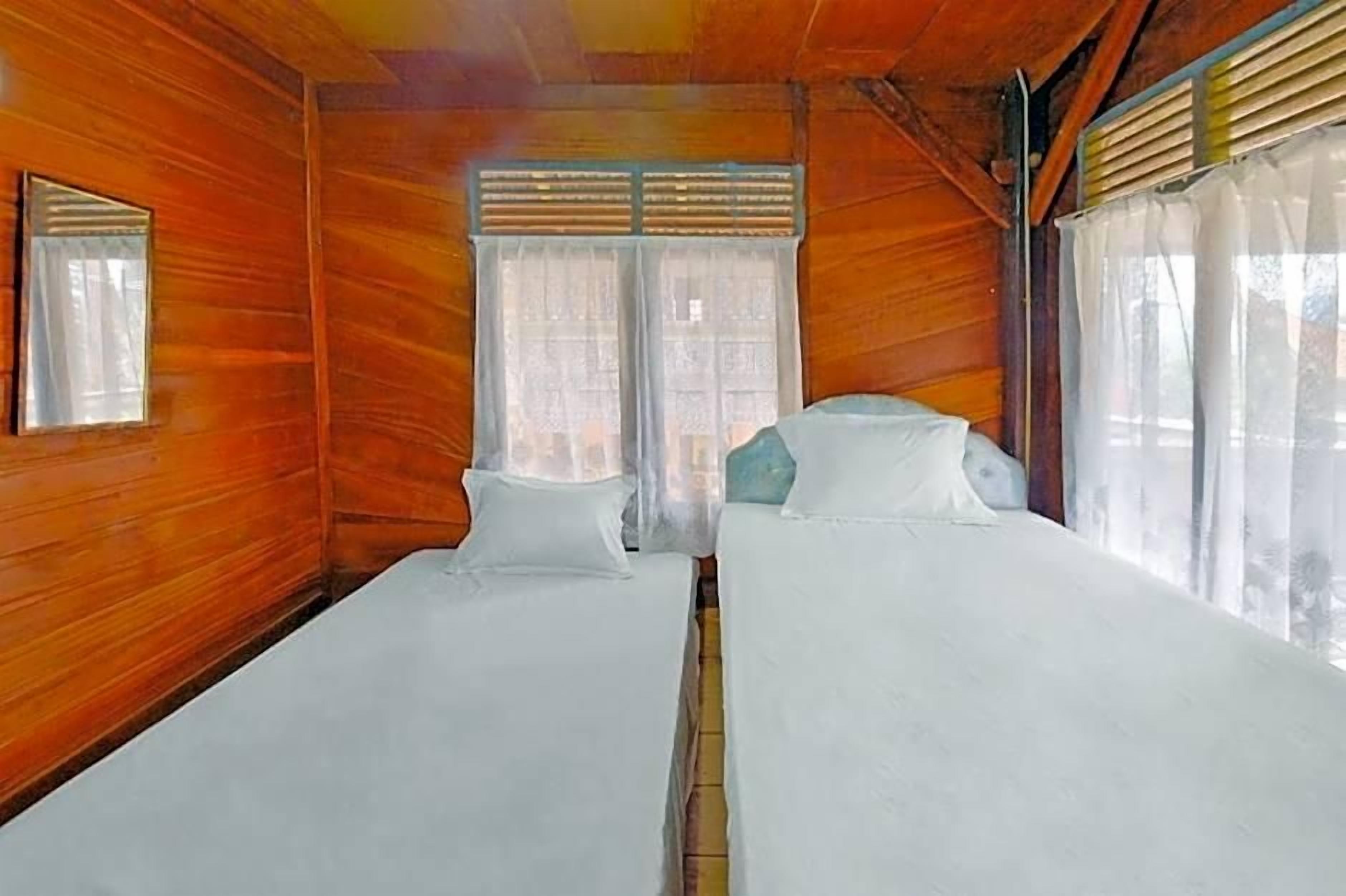 A room with wooden walls, two beds with white bedding, a window with curtains, and a small mirror on the wall.