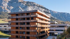 Ein mehrstöckiges Gebäude mit Holzfassade, vor einer gebirgigen Kulisse.