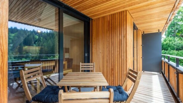 Eine Holzterrasse mit Tisch und Stühlen, eine Glastür, die auf Bäume blickt, und eine dunkle Wand.