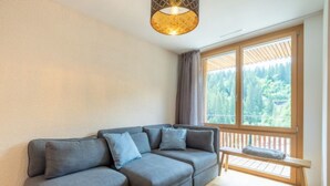 Ein modernes Wohnzimmer mit einem Ecksofa, einer Holzbank, einem großen Fenster mit Blick ins Grüne und einer Hängeleuchte.