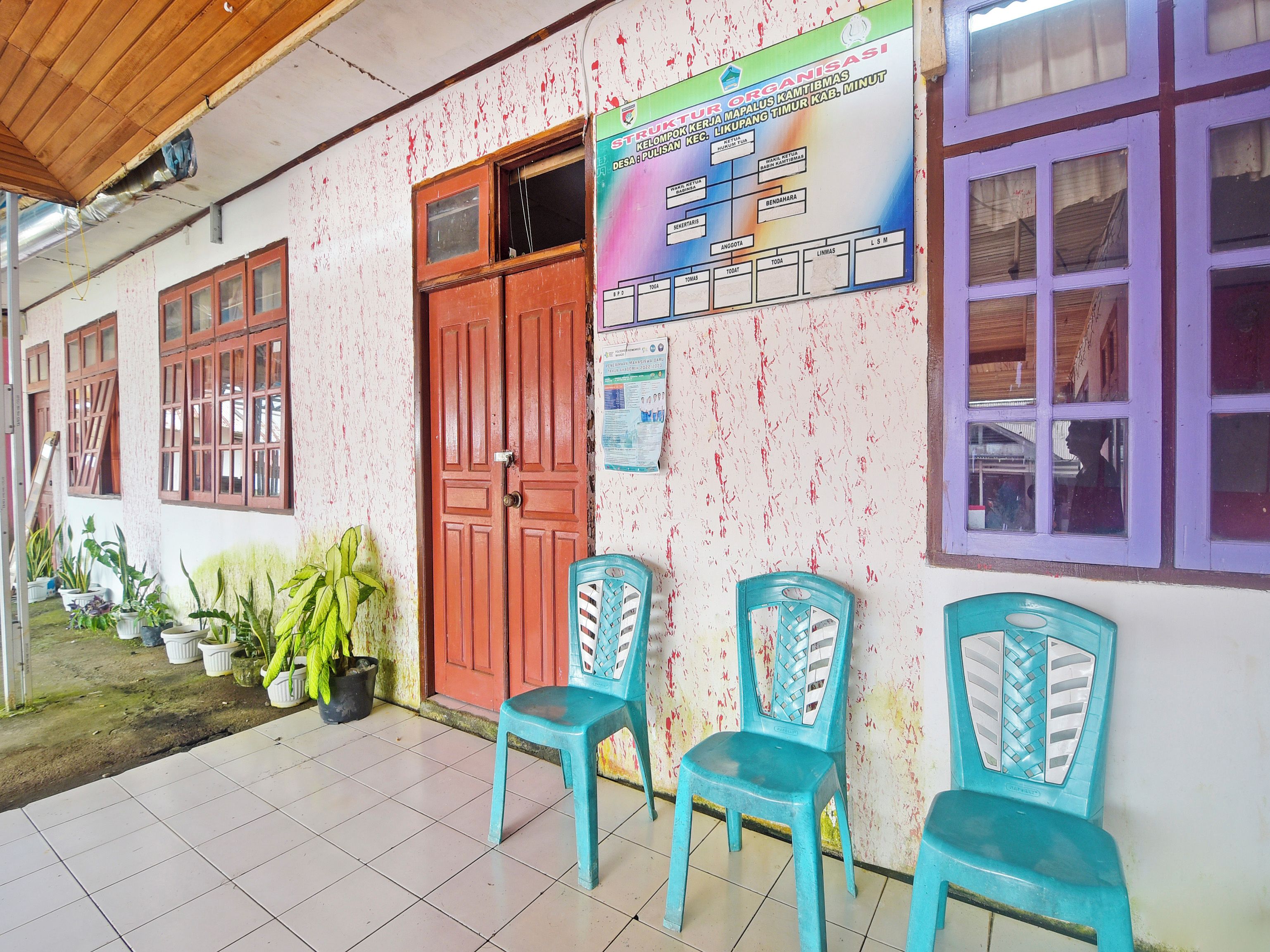 A building with a tiled floor, a wooden ceiling, and a door. There are potted plants and plastic chairs outside.