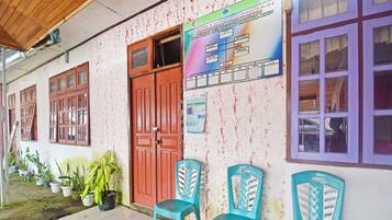 A building with a tiled floor, a wooden ceiling, and a door. There are potted plants and plastic chairs outside.