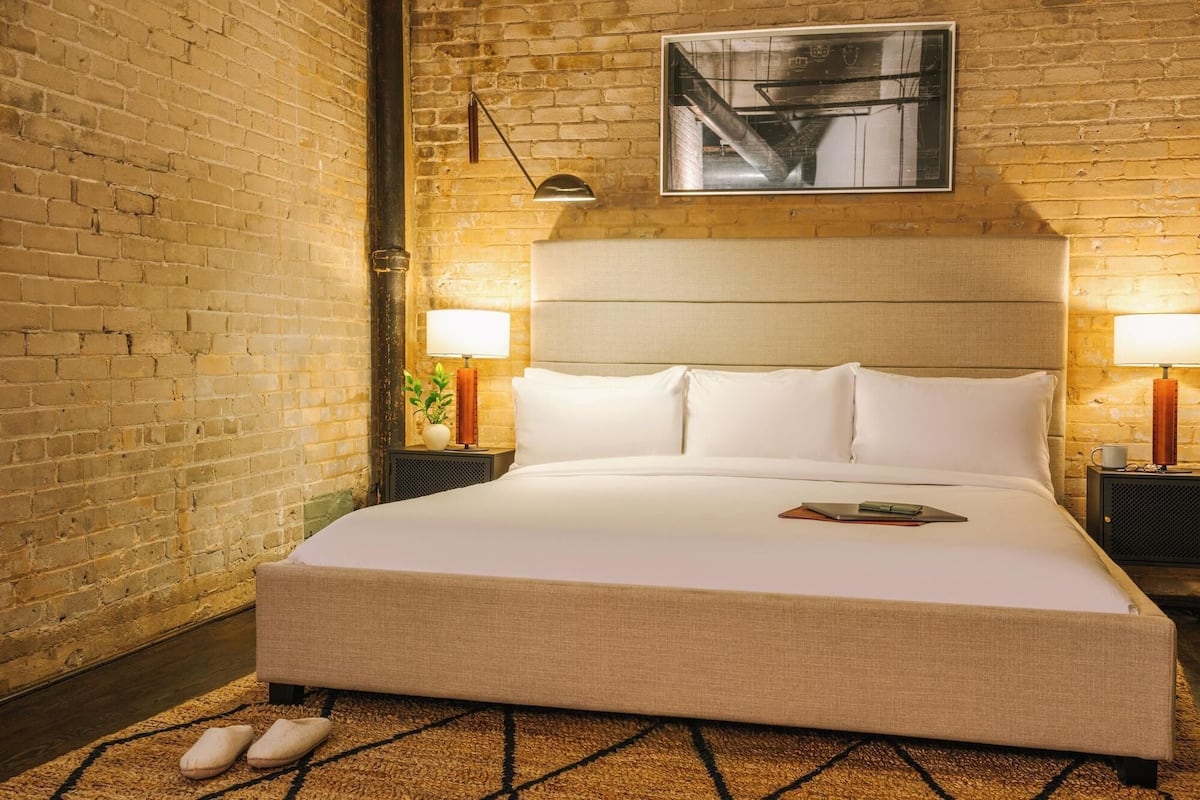 Ein Hotelzimmer mit Backsteinwand, einem großen Bett mit weißer Bettwäsche, zwei Nachttischen mit Lampen und einem gerahmten Bild an der Wand.