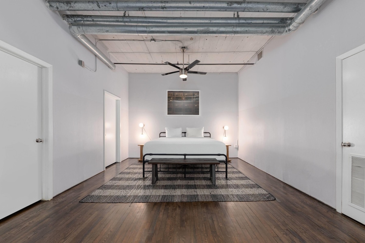 A bedroom with a bed, a ceiling fan, a light fixture, a wall-mounted artwork, and a wooden floor.