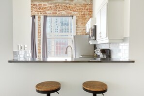 A kitchen with a brick wall, white cabinets, a window with curtains, and two stools. - Reside Houston Downtown A Wyndham Residence (Houston)