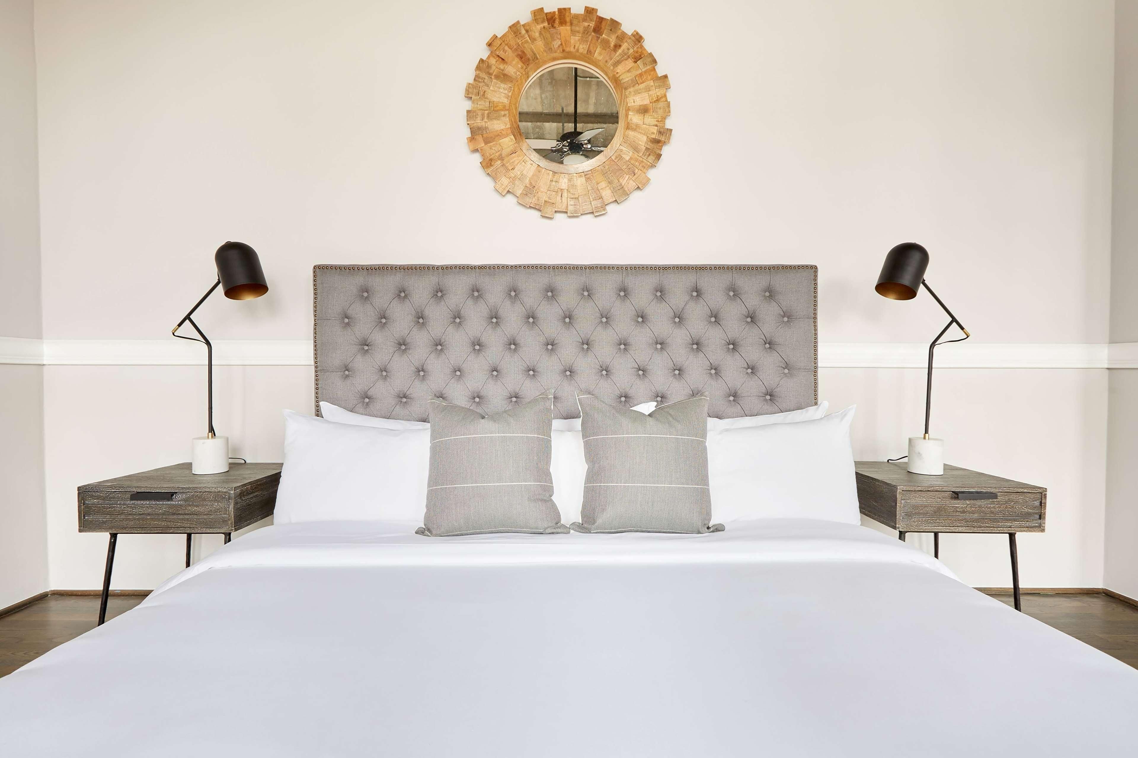 Habitación de hotel ordenada con cabecera capitonada, dos lámparas de noche, un espejo redondo y una cama con ropa de cama blanca.