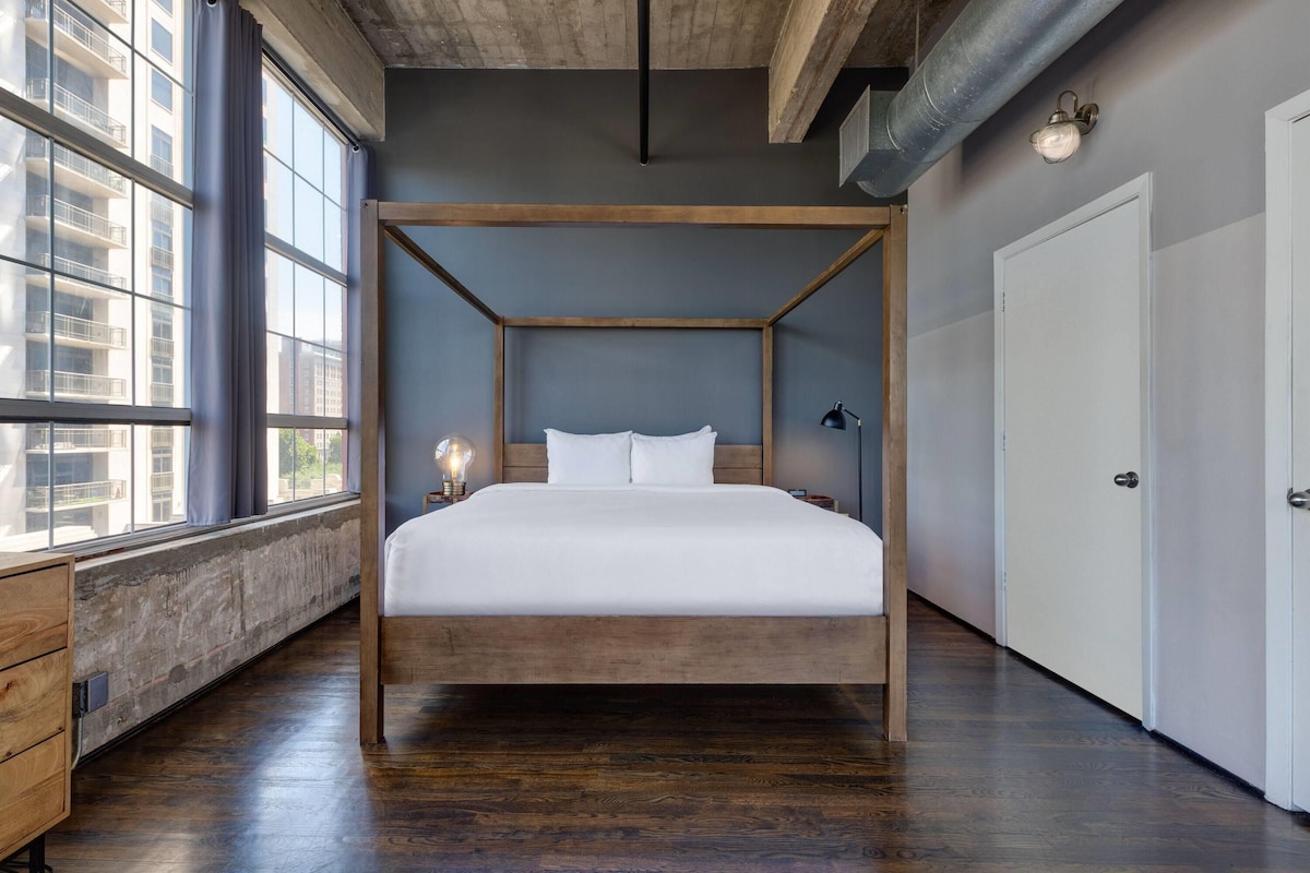 A four-poster bed in a room with large windows, a wooden floor, and a white door.