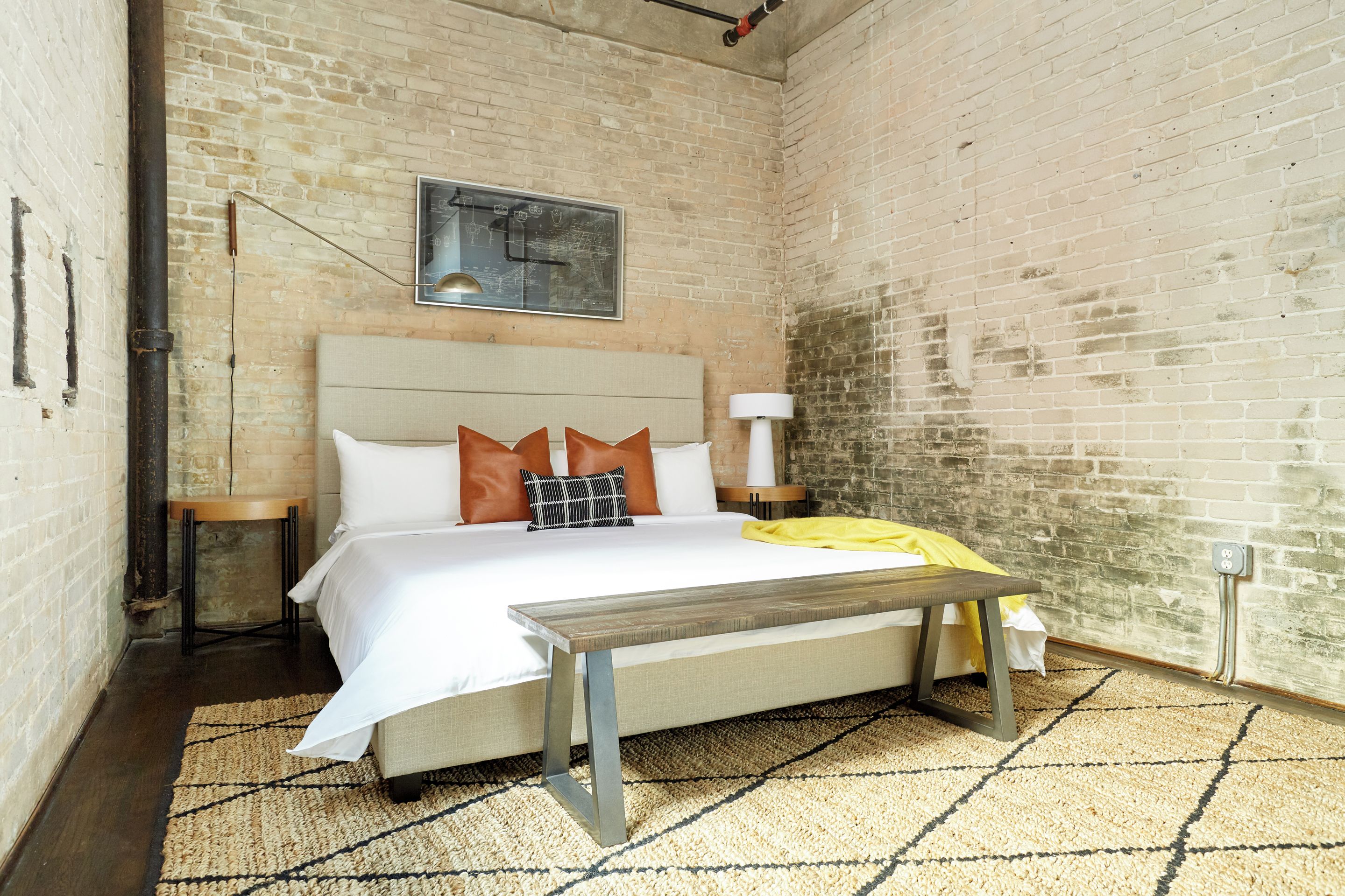 A bedroom with a bed, a bench, a bedside table, a lamp, and a brick wall.