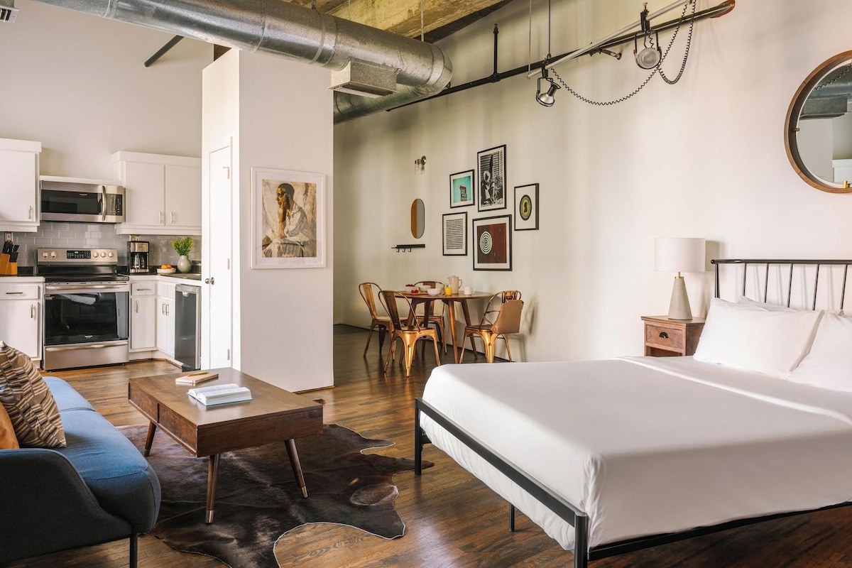 A modern hotel room with a bed, a desk, a dining area, and a kitchenette.