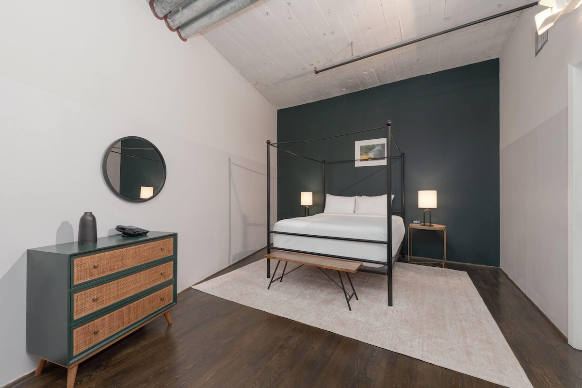 A bedroom with a four-poster bed, a dresser, a round mirror, and a bedside table with a lamp.