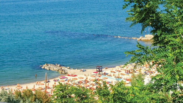 Een strand met parasols en ligbedden, een rotsachtige kustlijn en helderblauw water.