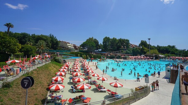 Ein großer Pool im Freien, umgeben von roten und weißen Sonnenschirmen, mit blauem Himmel darüber.