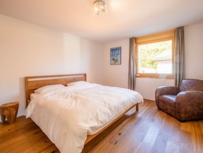 Ein Schlafzimmer mit einem hölzernen Bett, einem Sessel, einem kleinen Hocker, einem Fenster mit Vorhängen und einem an der Wand hängenden Bild.