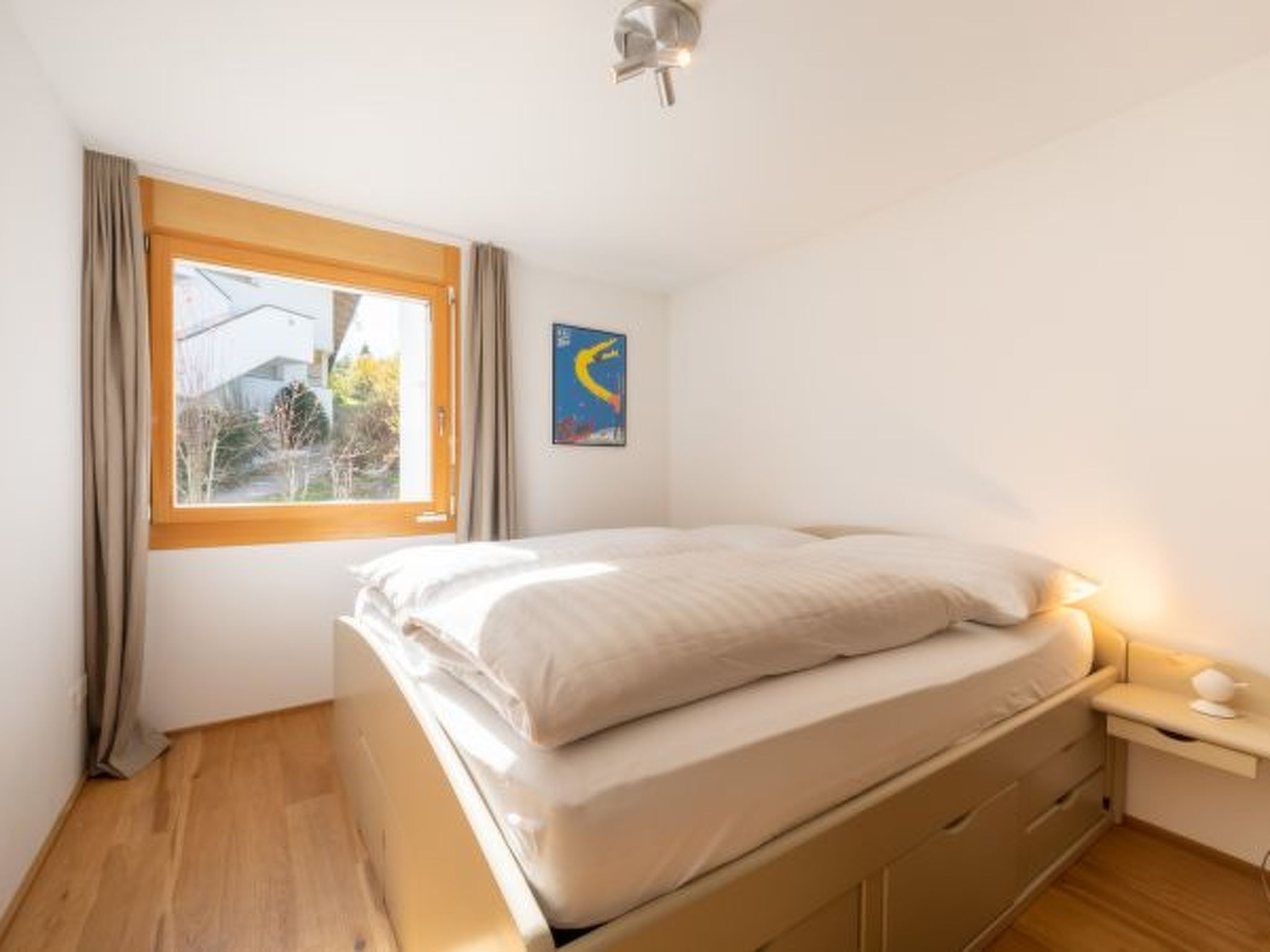Een slaapkamer met een bed, een nachtkastje, een raam met gordijnen en een aan de muur hangend kunstwerk.