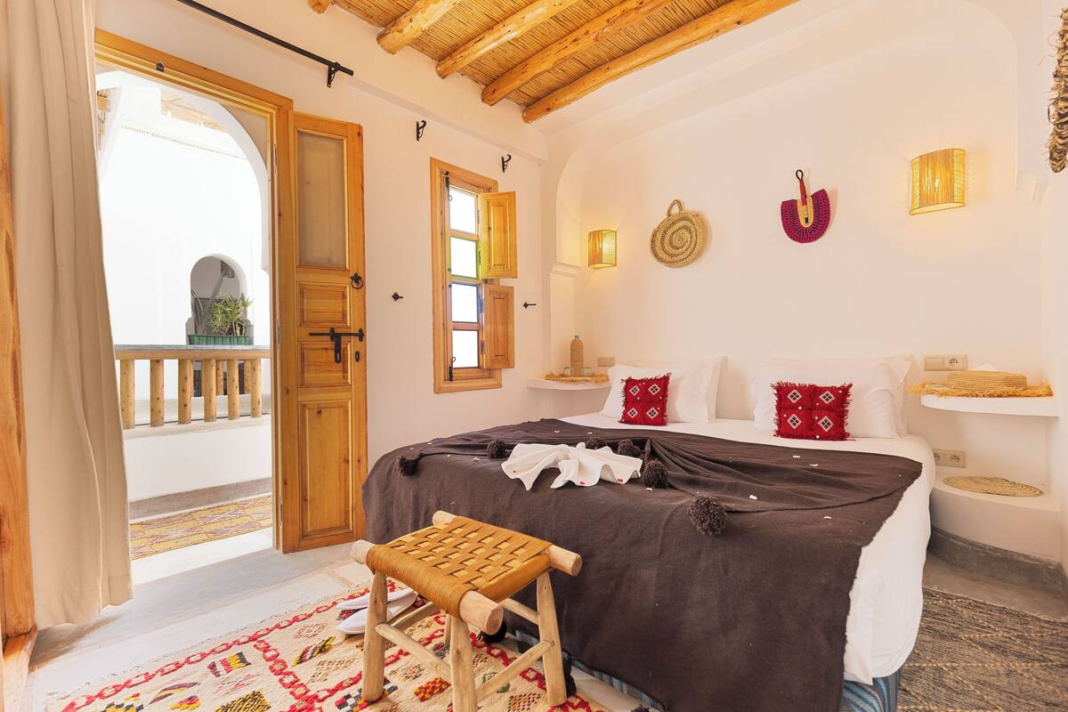 A bedroom with a bed, a wooden bench, a balcony, and a view of the outside.