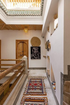 A hallway with a wooden railing, a patterned rug, a wooden door, and a wall-mounted artwork. - Riad Mama Square (Marrakech)