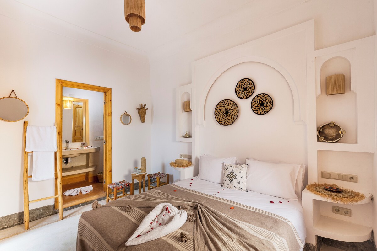 A well-lit, clean room with a bed, a wooden ladder, a mirror, a sink, and decorative wall art.