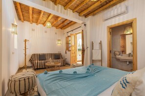 Una stanza con un letto, una poltrona, un tavolino, un appendiabiti e un bagno con uno specchio e un lavandino.