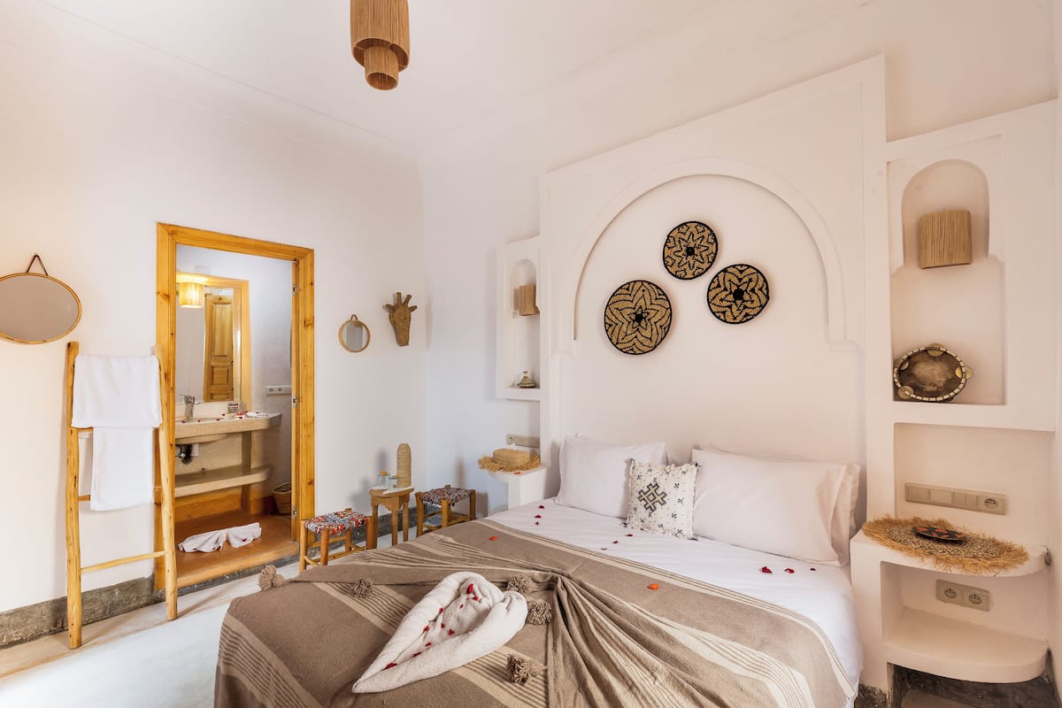 A well-lit, clean room with a bed, a wooden ladder, a mirror, a sink, and decorative wall art.