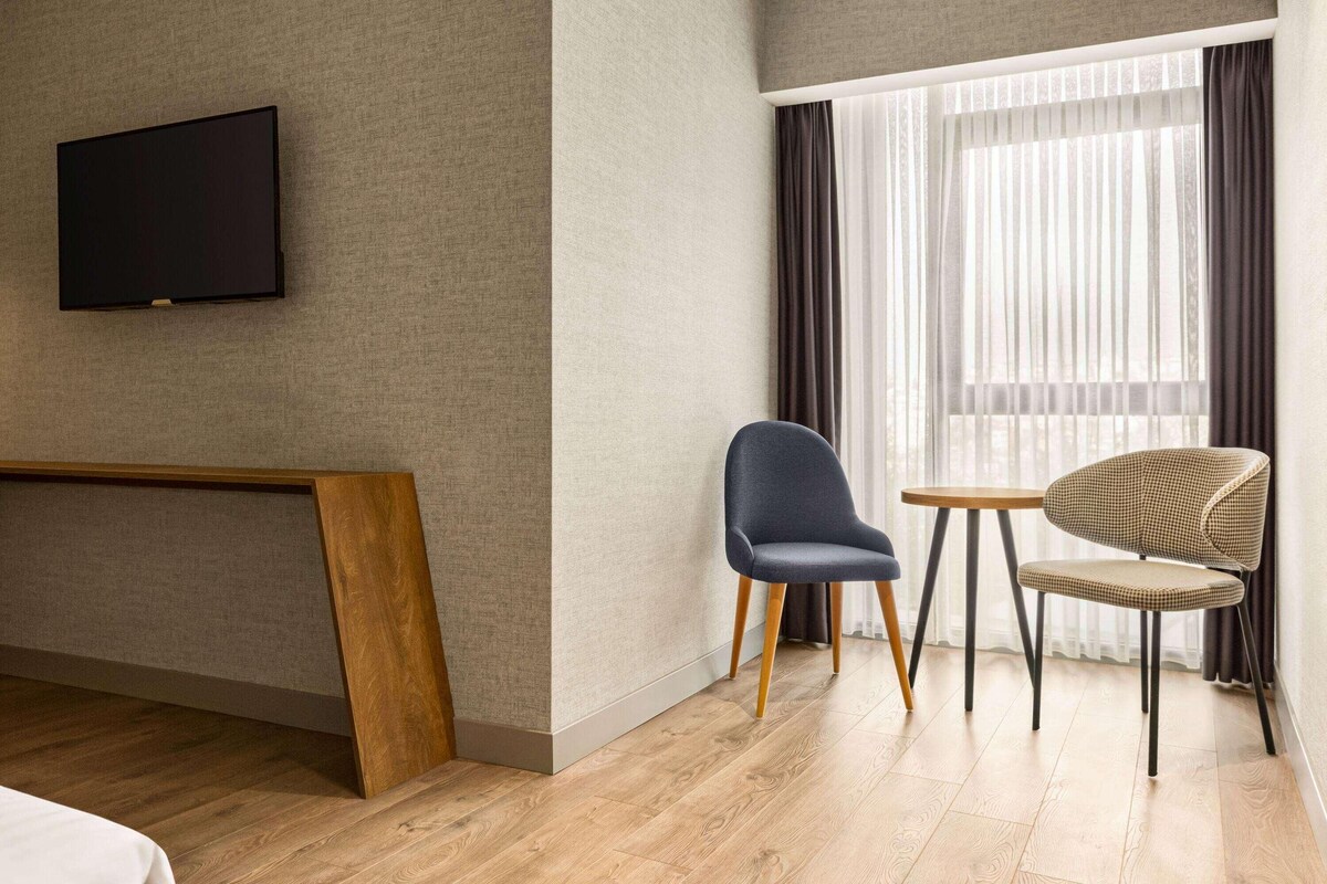 Habitación de hotel con una televisión de pantalla plana montada en la pared, un banco de madera, dos sillas y un ventanal con cortinas.