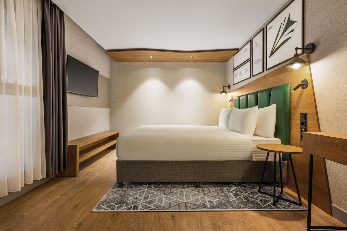 Una habitación de hotel moderna con una cama grande, un cabecero de madera, una mesita pequeña y un televisor montado en la pared.