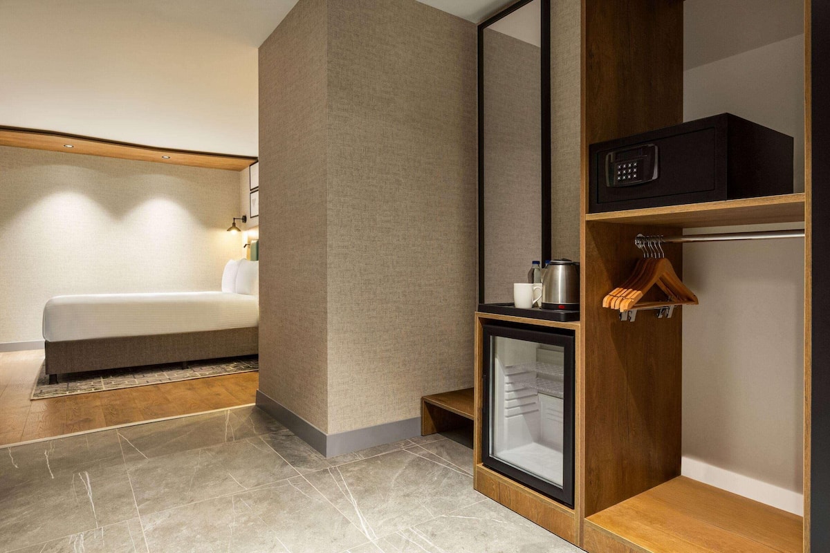 Ein modernes Hotelzimmer mit Bett, Minibar, kleinem Kühlschrank und einem Holzkabinett.