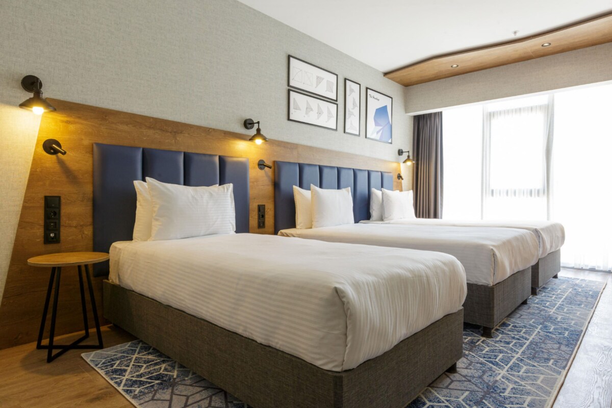 Habitación de hotel con dos camas, una mesita y vistas al exterior.