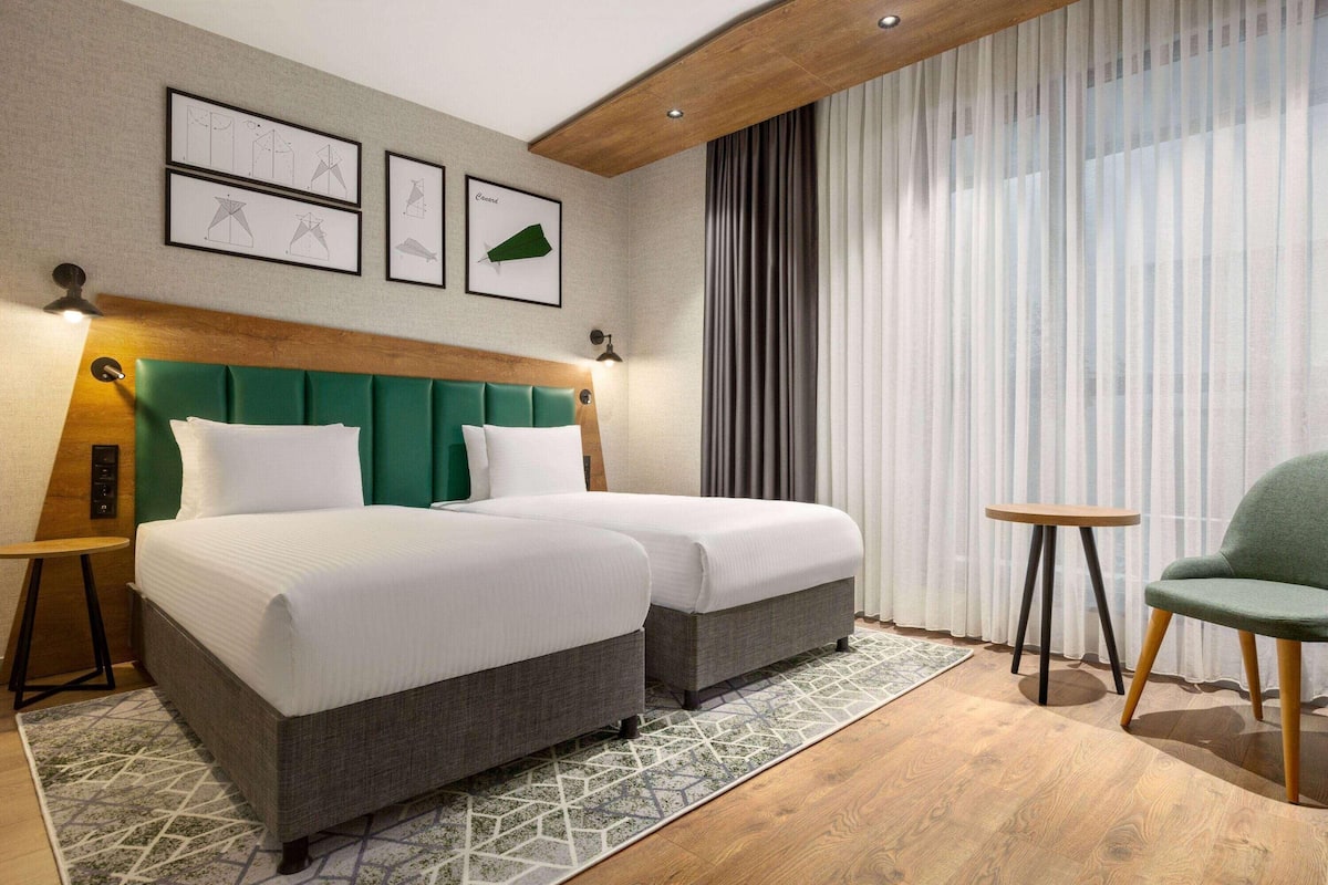 Ein Hotelzimmer mit zwei Betten, einem grünen gepolsterten Kopfteil, einem kleinen runden Tisch und einem Stuhl.