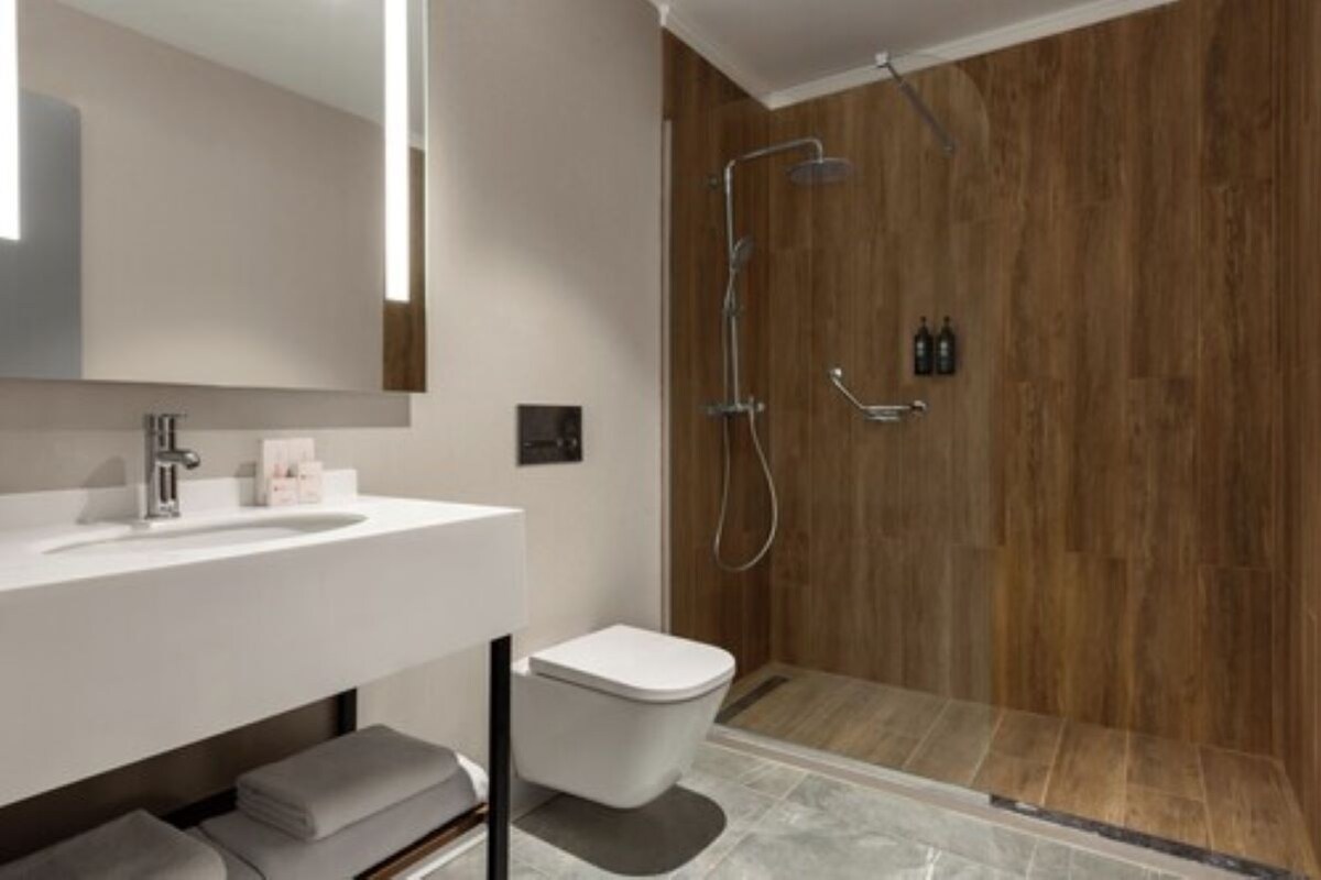Un baño moderno con lavamanos, espejo y retrete blancos, y una ducha con panel de madera.