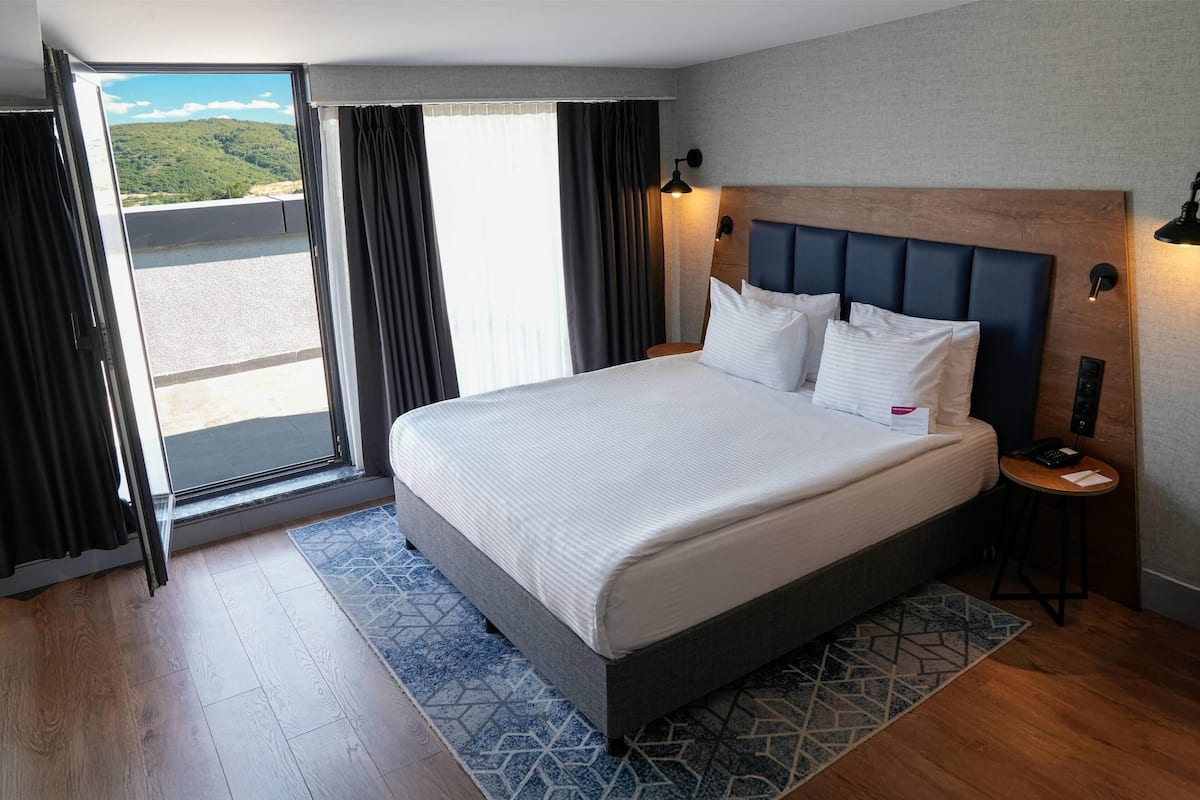 Ein Hotelzimmer mit einem großen Bett, einem Nachttisch, einem Fenster mit Vorhängen und Blick auf eine grüne Landschaft.