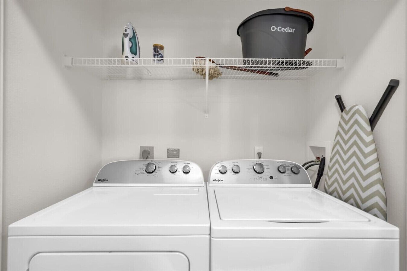 Uma área de lavanderia com duas máquinas de lavar Whirlpool brancas, uma prateleira com um balde preto O-Cedar e uma toalha estampada em padrão Chevron.