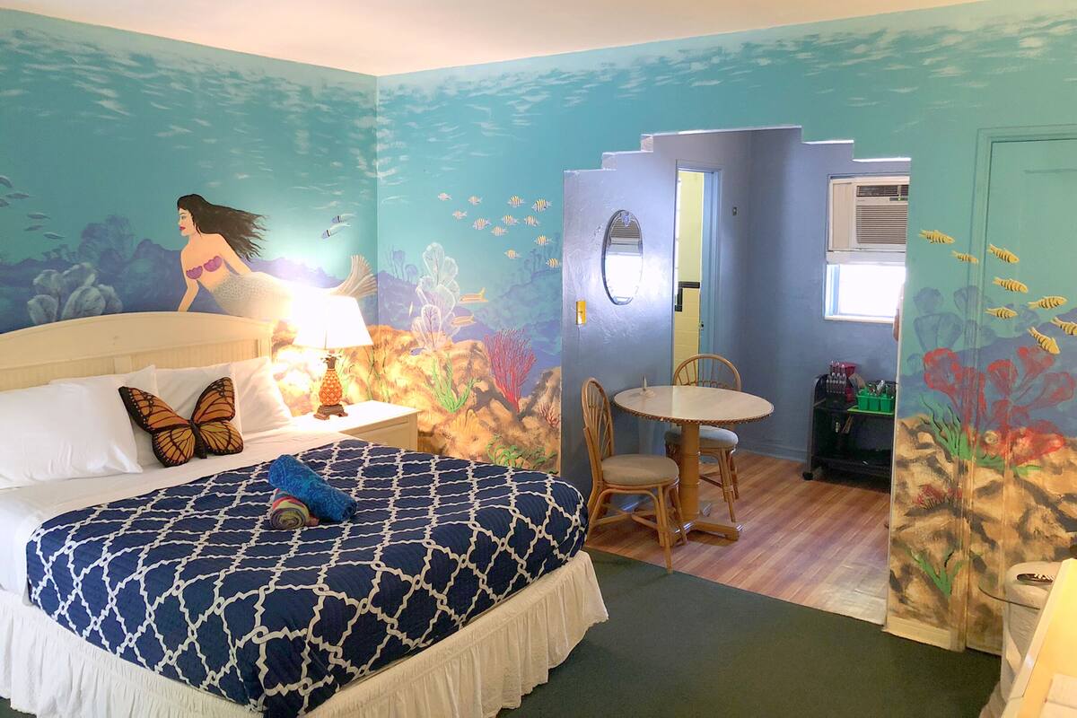 Habitación decorada con temática marina, que incluye una cama, una mesa, sillas y un mural con una sirena y peces.