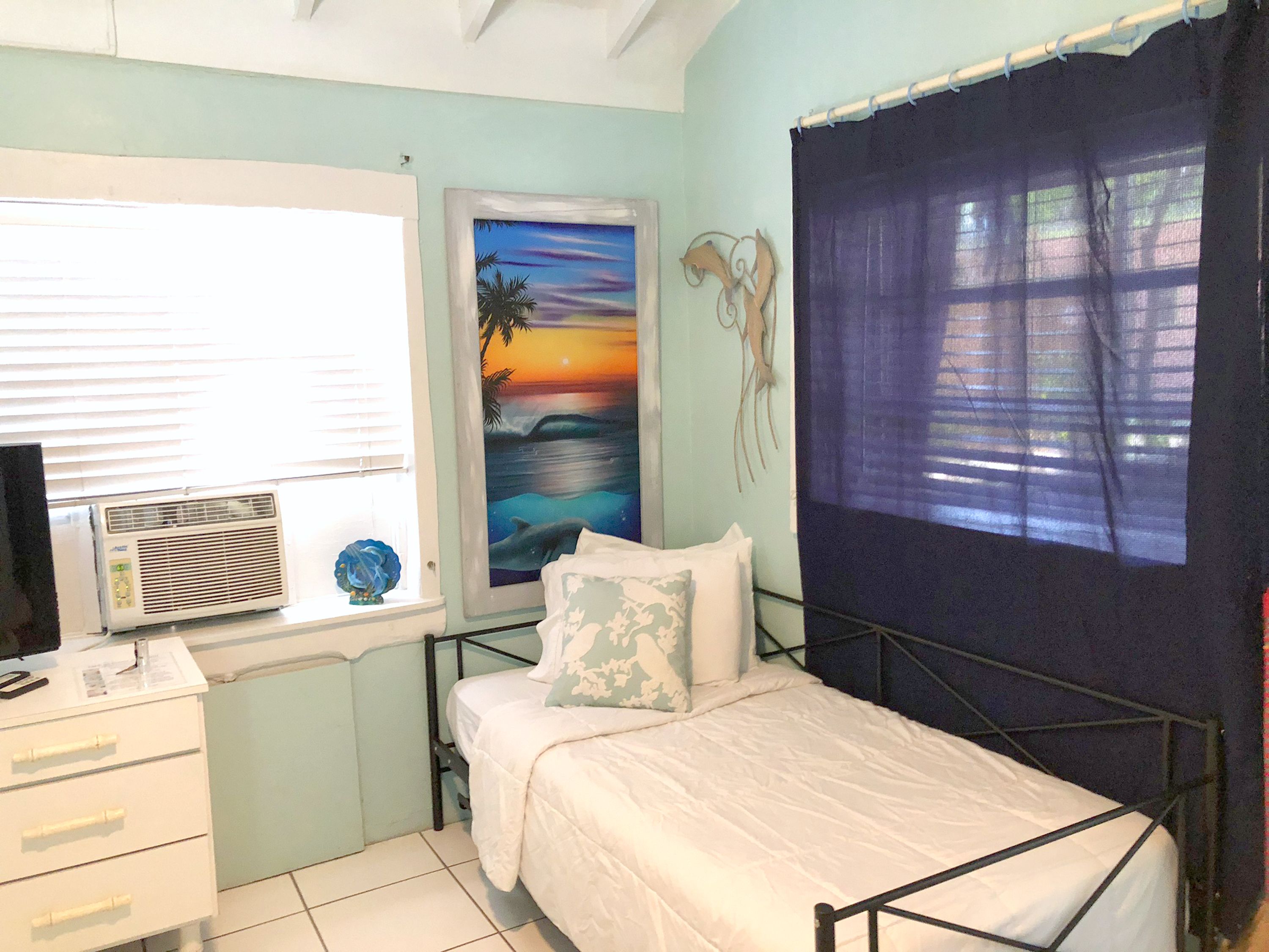 A bedroom with a bed, a desk, a window with blinds, a wall-mounted air conditioner, and a painting of a sunset.