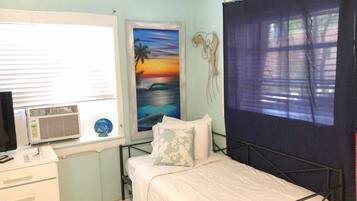 A bedroom with a bed, a desk, a window with blinds, a wall-mounted air conditioner, and a painting of a sunset.