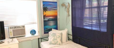 A bedroom with a bed, a desk, a window with blinds, a wall-mounted air conditioner, and a painting of a sunset.