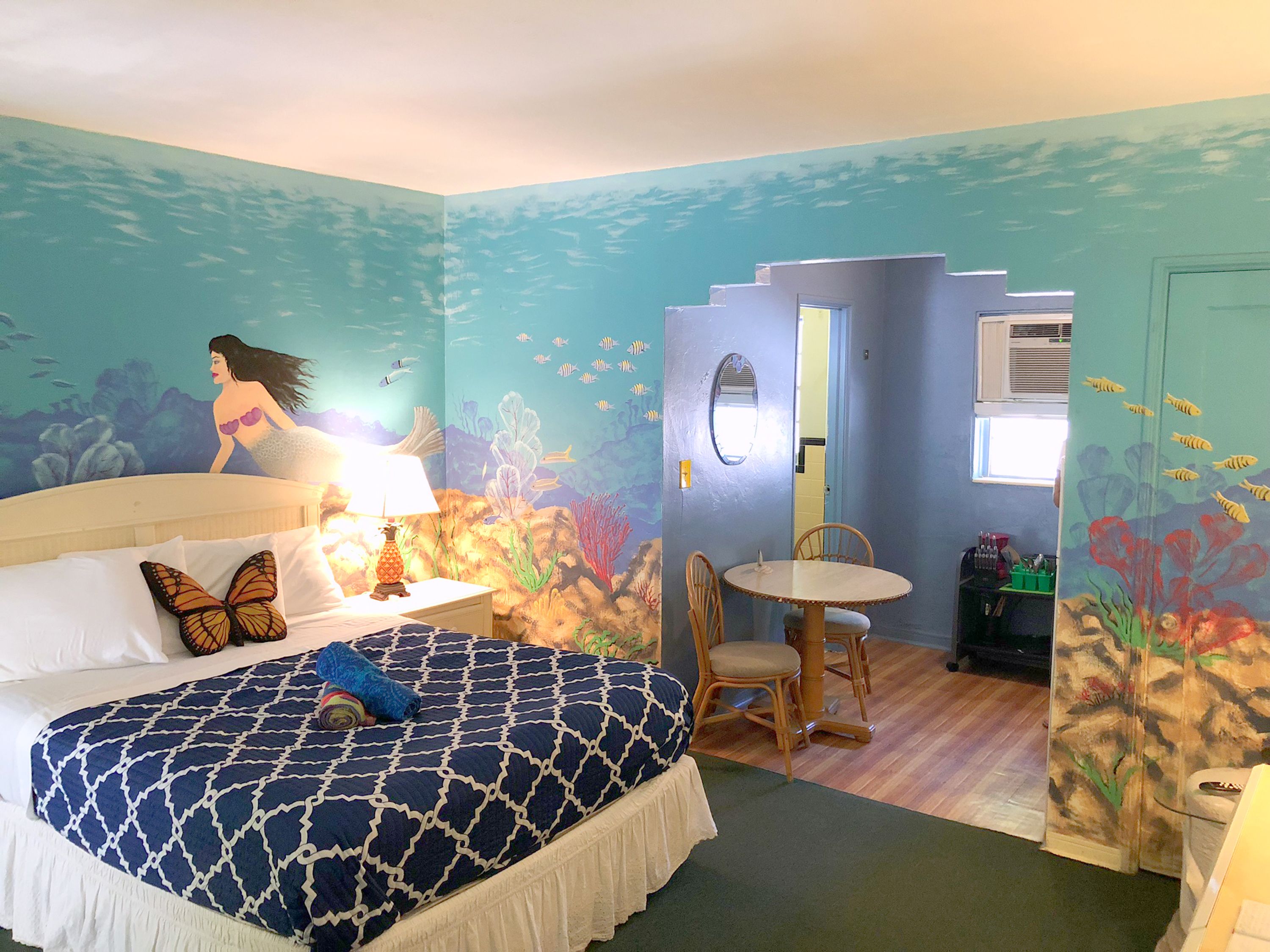 A room with a bed, a table, and chairs, decorated with an underwater theme including a mural of a mermaid and fish.