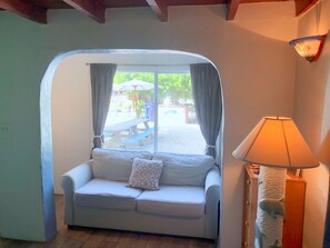 Habitación con sofá blanco, lámpara y vista a un espacio exterior con mesa de picnic y vegetación.