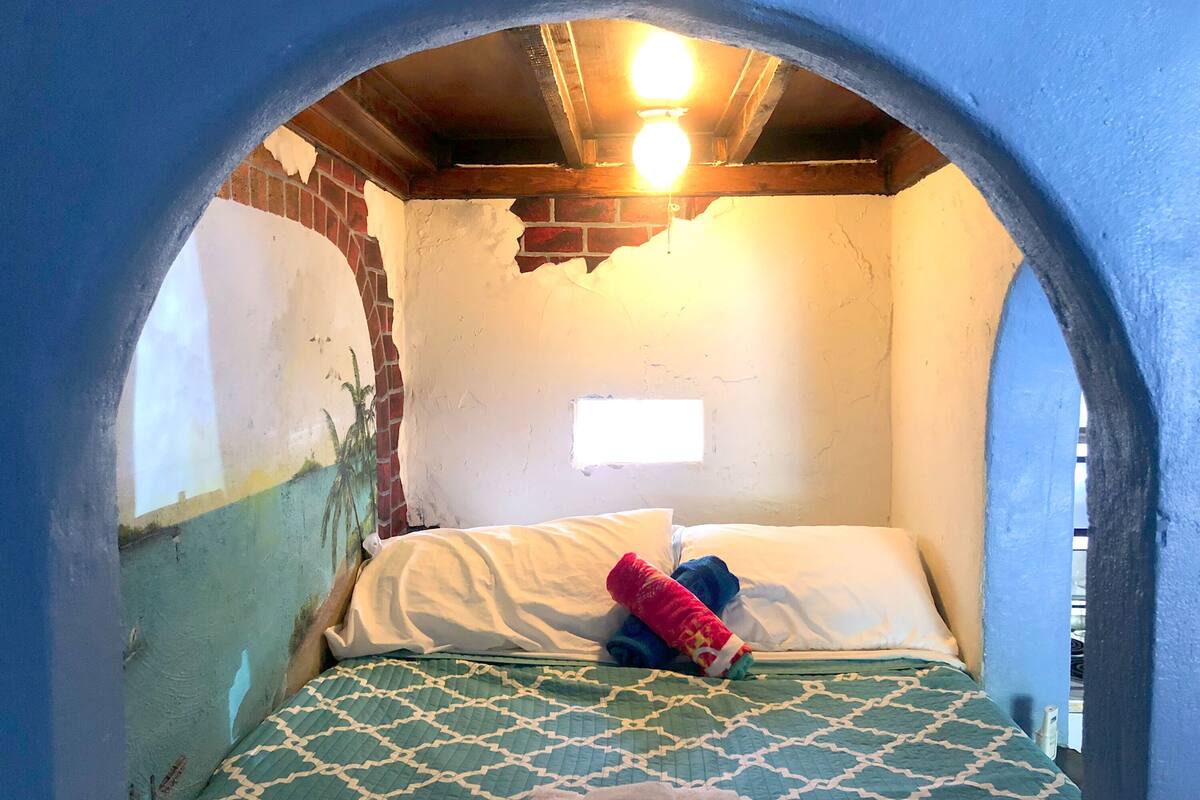 Una habitación con una cama, una ventana y una pared desconchada.