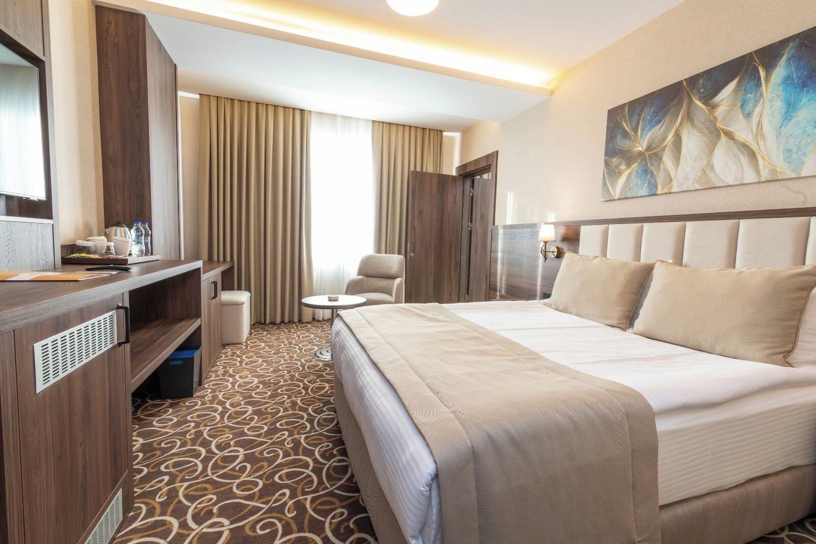 Quarto de hotel com cama, escrivaninha, cadeira, mesinha e um quadro na parede.