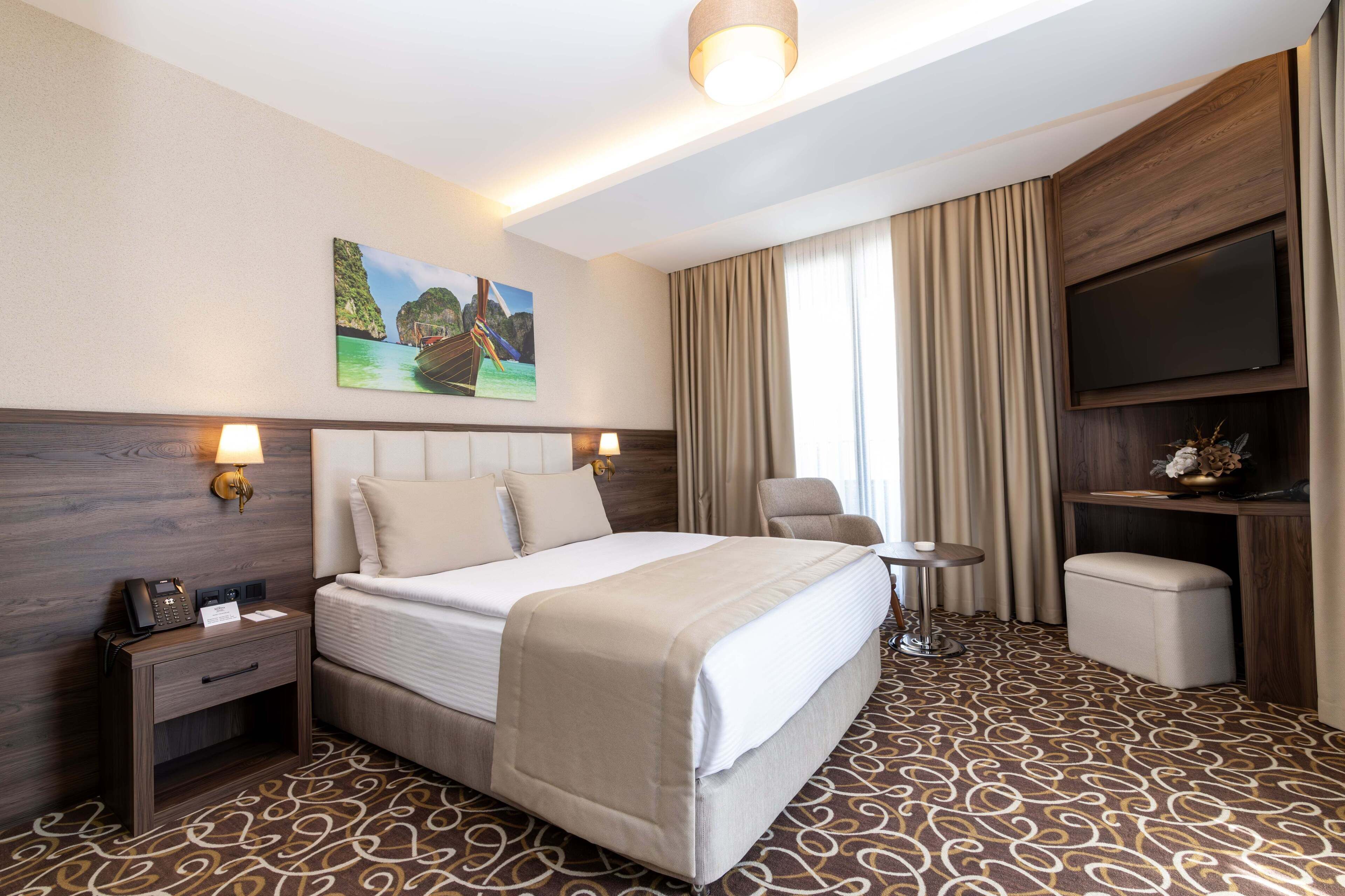 Quarto de hotel com cama, mesas de cabeceira, escrivaninha, cadeira, televisão e um quadro na parede.