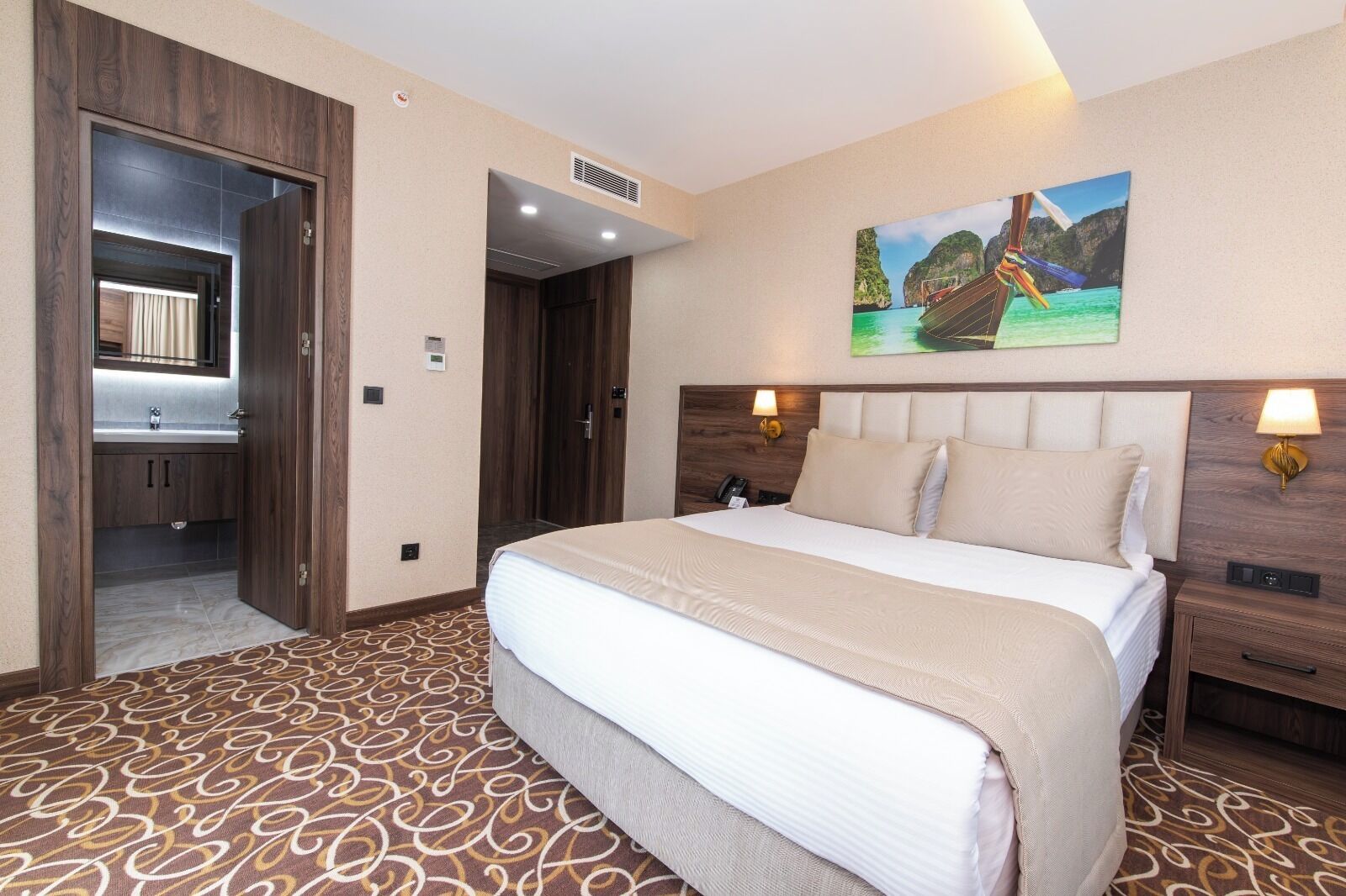 Quarto de hotel com uma cama grande, banheiro com espelho, pintura de um barco na parede e tapete estampado.