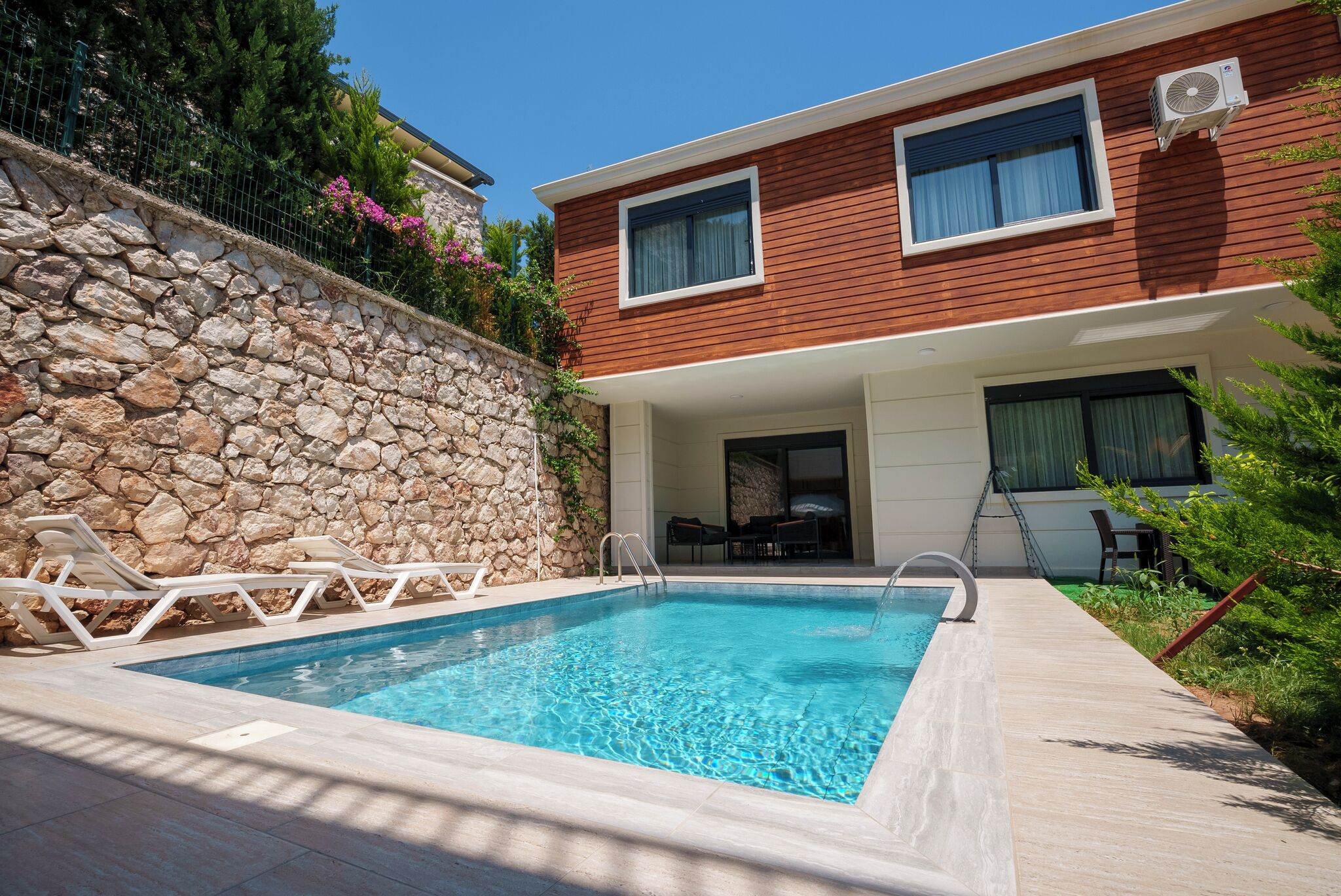 A swimming pool with lounge chairs, a stone wall, and a modern house with two windows and an air conditioning unit.